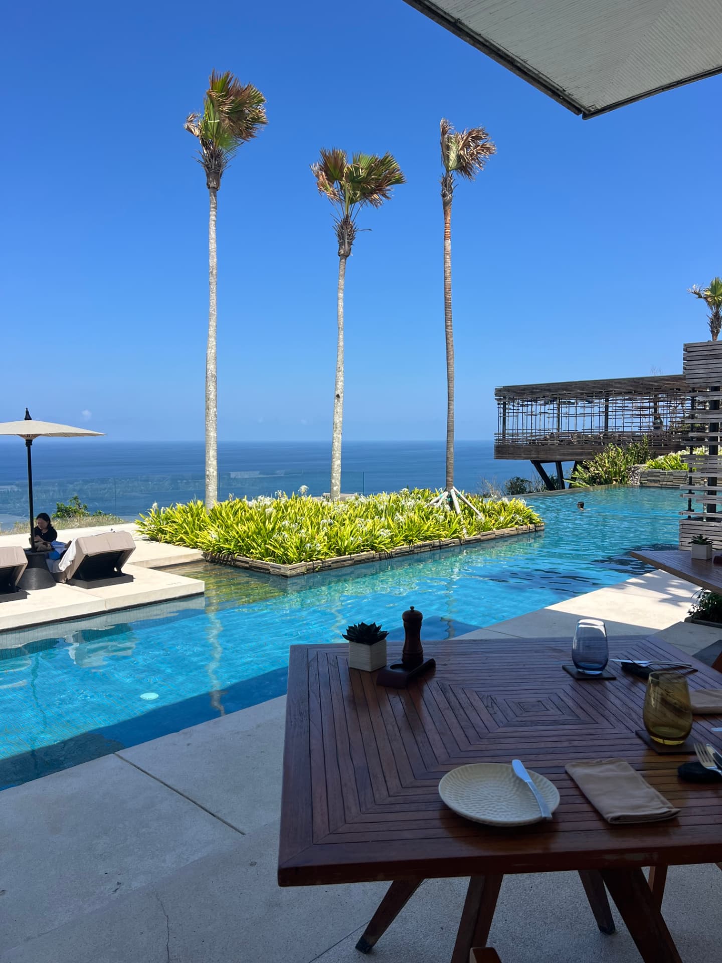A clifftop resort pool overlooking the ocean, with palms and a table set for dinner